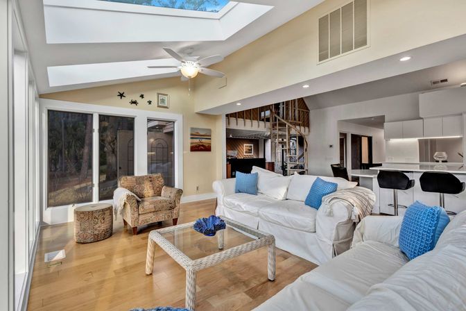 Bright, airy open-plan living room with skylights and ceiling fan, white sofas with blue pillows, wicker coffee table, patterned armchair, hardwood floors, glass sliding doors to a wooded yard, and a spiral staircase by a modern kitchen island with bar stools.