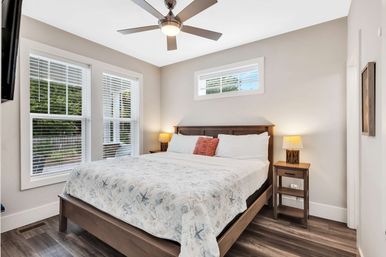 Sunlit modern bedroom with wood-frame bed dressed in a coastal-pattern quilt, matching wooden nightstands and lamps, ceiling fan, large white-trim windows and wood floors.