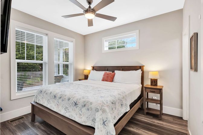 Sunlit modern bedroom with wood-frame bed dressed in a coastal-pattern quilt, matching wooden nightstands and lamps, ceiling fan, large white-trim windows and wood floors.