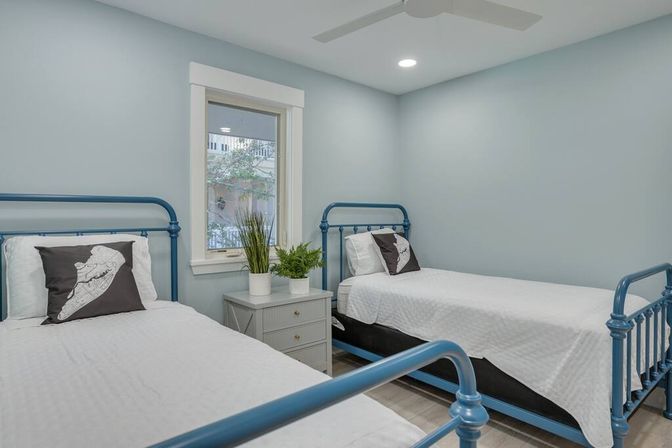 Coastal-style twin bedroom with two blue metal bed frames, crisp white bedding and map-print pillows, gray nightstand with potted plants, light blue walls, window and ceiling fan.