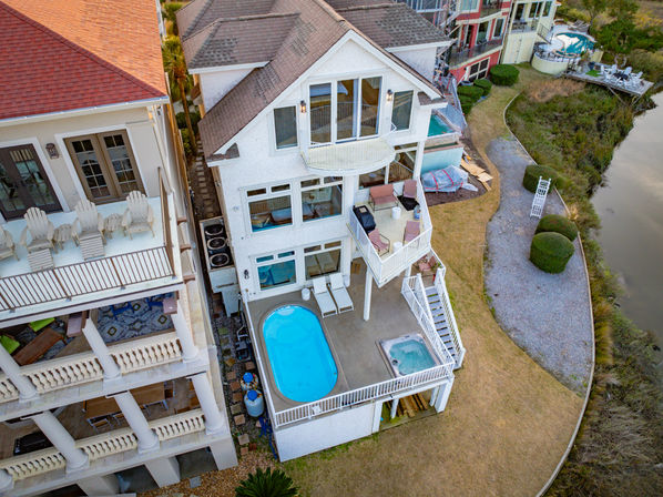 Aerial view of a multi-story waterfront beach house with balconies and patio seating, a small oval pool and hot tub on the lower deck, adjacent gravel path and marshy shoreline.