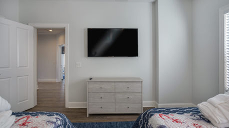 Bright modern guest bedroom with wall-mounted flat-screen TV above a light gray six-drawer dresser, hardwood floors, twin beds with blue nautical quilts, open door to hallway and window blinds.