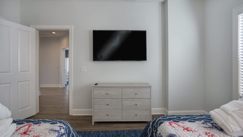 Bright modern guest bedroom with wall-mounted flat-screen TV above a light gray six-drawer dresser, hardwood floors, twin beds with blue nautical quilts, open door to hallway and window blinds.