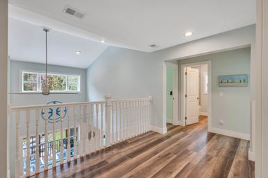 Bright upstairs landing in a coastal-inspired home with white spindle railing, wood-look plank floors, pale blue walls, pendant light and open doorway to bedroom and bathroom