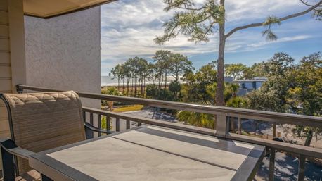 Sunlit beachfront balcony with patio table and chair overlooking coastal pine trees, sandy shoreline and distant ocean under a blue sky — relaxing ocean-view patio scene.