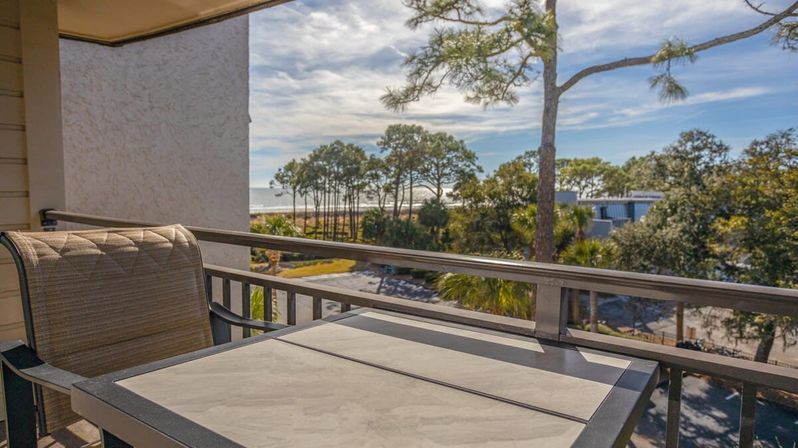 Sunlit beachfront balcony with patio table and chair overlooking coastal pine trees, sandy shoreline and distant ocean under a blue sky — relaxing ocean-view patio scene.