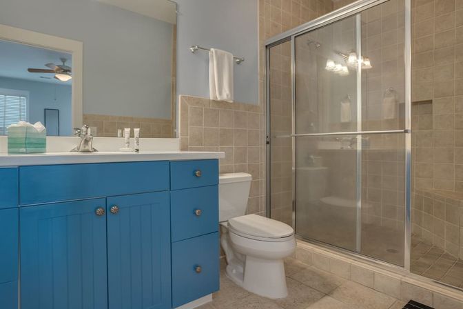 Modern bathroom with a cheerful blue vanity and white countertop, toilet, glass sliding shower, beige tiled walls and floor, and a white towel on a bar.