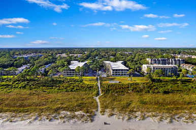 Aerial view of beachfront condos and low-rise buildings behind dune grasses, with a wooden boardwalk path leading from a wide sandy beach through coastal vegetation to landscaped grounds under a bright blue sky.