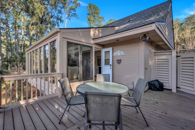 Cozy backyard wood deck patio with a round glass table and four mesh chairs, attached beige sunroom with large windows and white door, surrounded by tall pine trees under a clear blue sky.