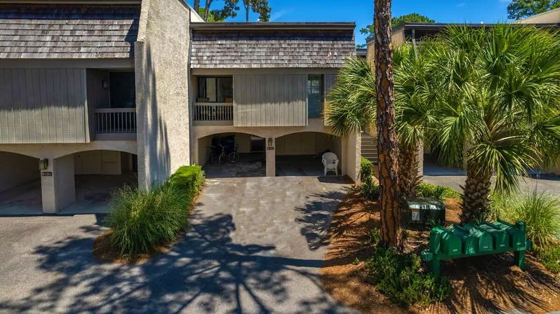 Sunny coastal condominium carport with bicycles, a white plastic chair, palm trees and green cluster mailboxes.