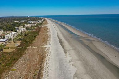 Aerial view of a long sandy beach and calm blue ocean along a barrier island coastline, with coastal dunes and beachfront condos lining the shore under a clear sky.