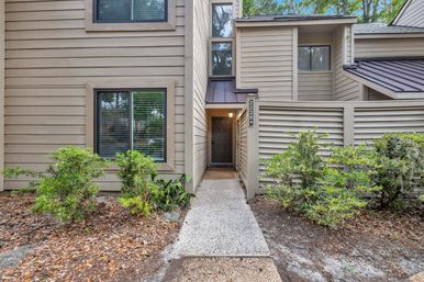 Pebble walkway leading to a recessed dark front door of a beige wood‑sided townhouse, flanked by horizontal privacy fencing, windows with blinds, and low green shrubs.