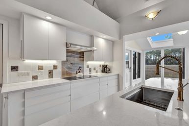 Sunlit modern white kitchen with quartz countertops, built-in induction cooktop and coastal tile backsplash, brass spring pull-down faucet over stainless sink, skylight and glass doors framing a marsh and pine-tree view