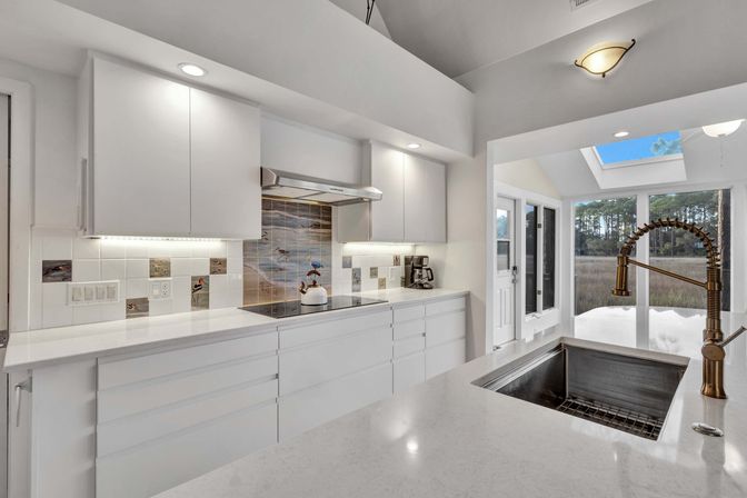 Sunlit modern white kitchen with quartz countertops, built-in induction cooktop and coastal tile backsplash, brass spring pull-down faucet over stainless sink, skylight and glass doors framing a marsh and pine-tree view