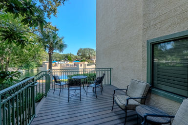 Cozy lakeside balcony with cushioned patio chairs and a metal bistro table on a wood deck, green railing framing a view of palm trees, a pool area with umbrellas and a bright blue sky.