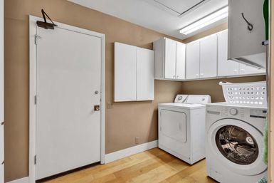 Home laundry room with front-load washer, top-load dryer, white cabinets and laundry basket against beige walls, hardwood floor, fluorescent ceiling light and white utility door
