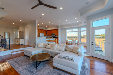 Open-concept waterfront living room in a modern coastal home with a cream L-shaped sectional, round brass coffee table, hardwood floors, wood-cabinet kitchen, dining nook by large windows and a balcony door overlooking a marsh view.