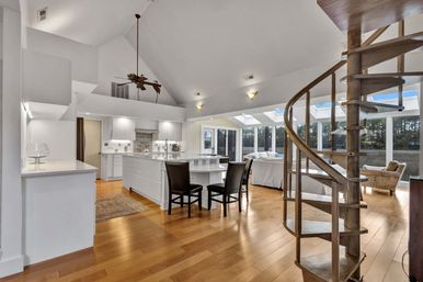 Sunlit open-concept kitchen and living area with vaulted ceilings and skylights, white island with bar stools, warm hardwood floors, palm-blade ceiling fan and a wooden spiral staircase overlooking marsh and trees through floor-to-ceiling windows.