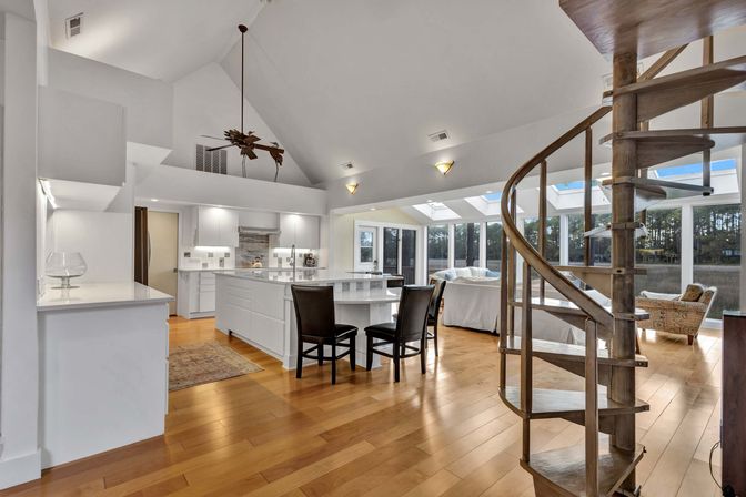Sunlit open-concept kitchen and living area with vaulted ceilings and skylights, white island with bar stools, warm hardwood floors, palm-blade ceiling fan and a wooden spiral staircase overlooking marsh and trees through floor-to-ceiling windows.