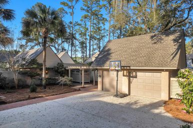 Sunny suburban driveway with a two-car garage and portable basketball hoop, shingled roof and carport, framed by palm trees, tall pines and landscaped beds under a bright blue sky.