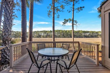 Inviting wooden deck patio with round glass table and four chairs overlooking a coastal marsh framed by palm and pine trees under a bright blue sky.