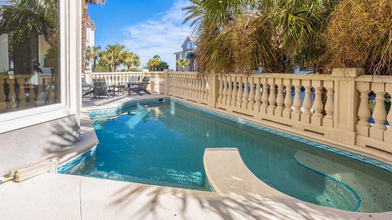 Sunny coastal balcony plunge pool with classical balustrade, palm trees and lounge chairs in a seaside neighborhood.