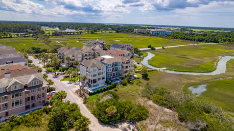 Aerial drone view of colorful coastal townhouses and palm-lined streets overlooking a winding tidal creek and green salt marsh under a partly cloudy sky