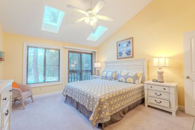 Sunny master bedroom with vaulted ceiling and skylights, white ceiling fan, king bed with yellow-gray daisy quilt, white nightstands and lamps, sliding glass door to a deck with a leafy view.