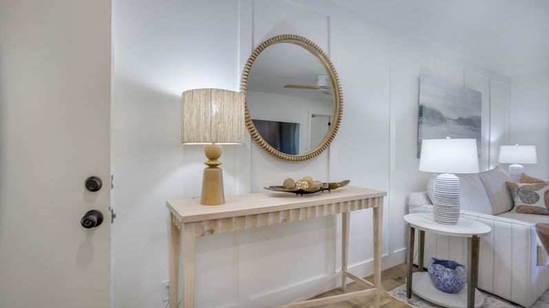 Bright coastal-inspired entryway with light wood console table, round woven mirror, textured lamp and decorative bowl, opening into a neutral living room with striped sofa and white table lamps
