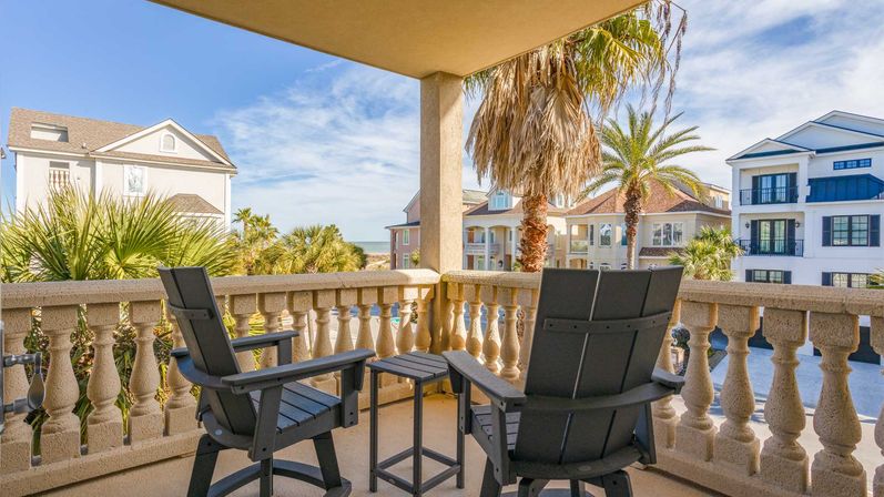 Coastal balcony with two black chairs and small table, stone balustrade, palm trees and a distant ocean view past beach houses