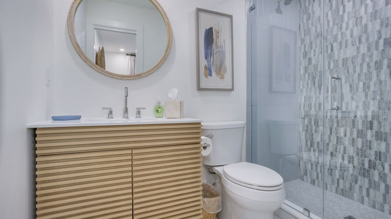 Bright modern bathroom with round mirror, ribbed light-wood vanity, white sink and chrome faucet, toilet, abstract wall art, and glass-enclosed shower with gray mosaic tile.