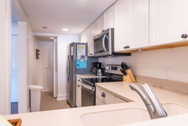 Bright modern galley kitchen with stainless steel refrigerator, microwave and stove, white cabinets, beige countertops, sink and faucet in foreground, knife block and coffee maker on the counter