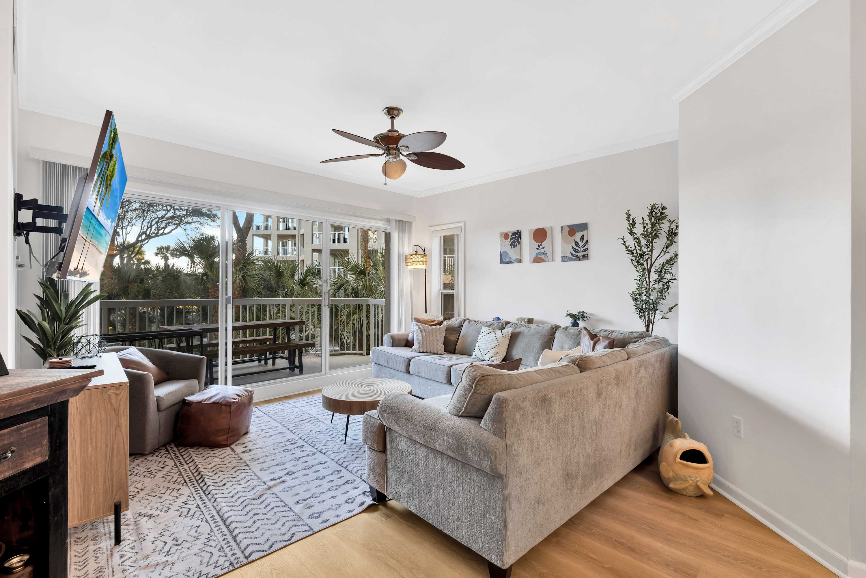 Bright beachside condo living room with a gray sectional, patterned rug and round coffee table, ceiling fan and wall-mounted TV, sliding glass doors to a balcony overlooking palm trees and nearby buildings, light wood floors and potted plant.