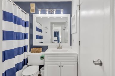 Bright nautical-style residential bathroom with navy-and-white striped shower curtain, white vanity and sink with chrome faucet, large framed mirror, toilet topped with a wicker basket, and modern vanity lights.