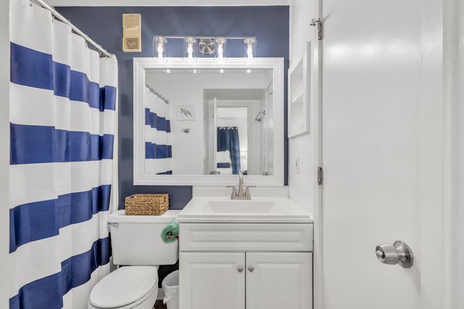 Bright nautical-style residential bathroom with navy-and-white striped shower curtain, white vanity and sink with chrome faucet, large framed mirror, toilet topped with a wicker basket, and modern vanity lights.