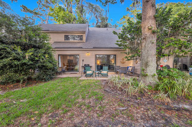Cozy two-story townhouse backyard patio with sliding glass doors, Adirondack chairs and outdoor dining set under an oak tree draped in Spanish moss, grassy lawn in foreground.