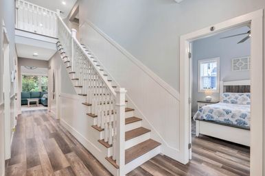 Bright modern entryway with white wooden staircase and wide-plank hardwood floors, hallway leading to a sunlit living room and a coastal-themed bedroom with coral-print bedding.