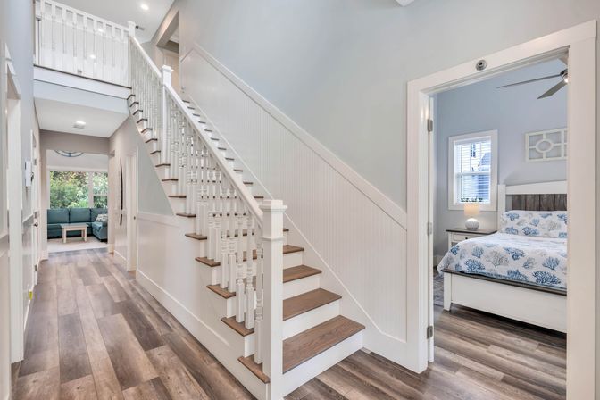 Bright modern entryway with white wooden staircase and wide-plank hardwood floors, hallway leading to a sunlit living room and a coastal-themed bedroom with coral-print bedding.