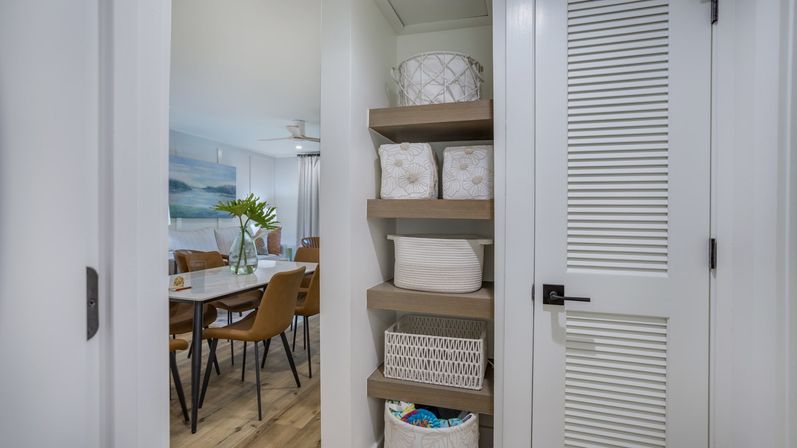Built-in wooden shelves with woven baskets beside a white louvered door, opening to a modern dining area with a marble table, tan chairs, glass vase of green leaves and light wood floors