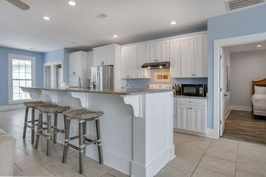 Open-concept coastal-style kitchen with light blue walls, white shaker cabinets, granite breakfast bar with three woven wooden bar stools, stainless refrigerator and range hood, microwave on tiled floor and doorway to a bedroom.