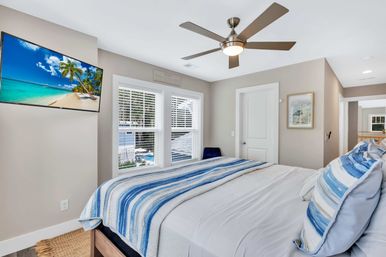 Bright coastal-style master bedroom with king bed in blue striped bedding, modern ceiling fan, wall-mounted TV showing a tropical beach, and double windows with white blinds.