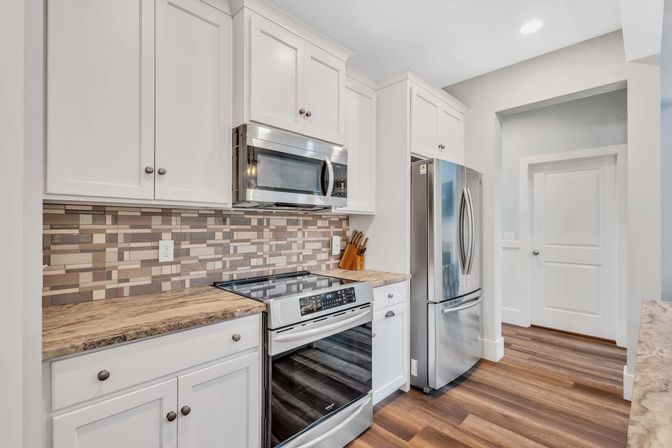 Bright modern kitchen with white cabinets, stainless steel fridge, microwave and range, neutral mosaic tile backsplash, wood-look countertops and warm plank flooring.