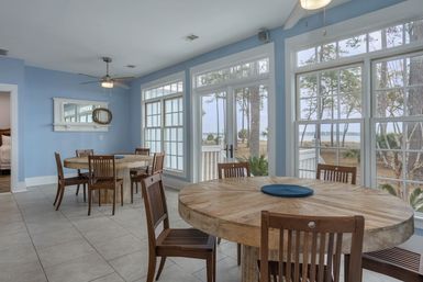 Bright coastal dining room with light-blue walls, round reclaimed-wood tables and wooden chairs, tile floor and ceiling fans, and large windows/French doors framing a bayfront view with trees.