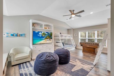 Bright beach-themed family game room loft with white bunk beds, built-in TV showing a tropical beach, two navy bean bags, starfish rug, foosball table and large window