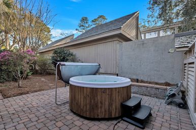 Round wooden outdoor hot tub with white rim and black steps on a paver backyard patio, cover lifter and hose reel nearby, bordered by a privacy fence, shrubs and flowering bushes under a blue sky