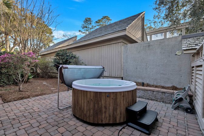 Round wooden outdoor hot tub with white rim and black steps on a paver backyard patio, cover lifter and hose reel nearby, bordered by a privacy fence, shrubs and flowering bushes under a blue sky