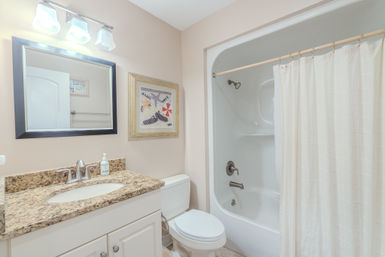 Bright coastal-themed bathroom with granite countertop vanity and stainless faucet, framed mirror, white toilet, and tub-shower with beige fabric curtain against soft beige walls.