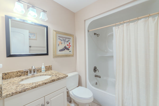 Bright coastal-themed bathroom with granite countertop vanity and stainless faucet, framed mirror, white toilet, and tub-shower with beige fabric curtain against soft beige walls.