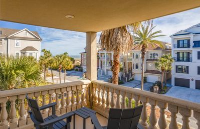 Sunny coastal balcony with two dark Adirondack chairs, stone balustrade, palm trees and pastel beach houses with ocean view in the distance.