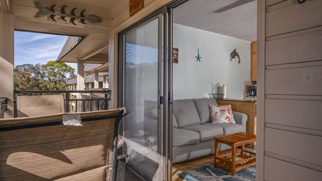 Balcony of a coastal condo with outdoor seating and sliding glass door opening to a bright living room with gray sofa, nautical wall decor, wooden coffee table, and tree-lined view